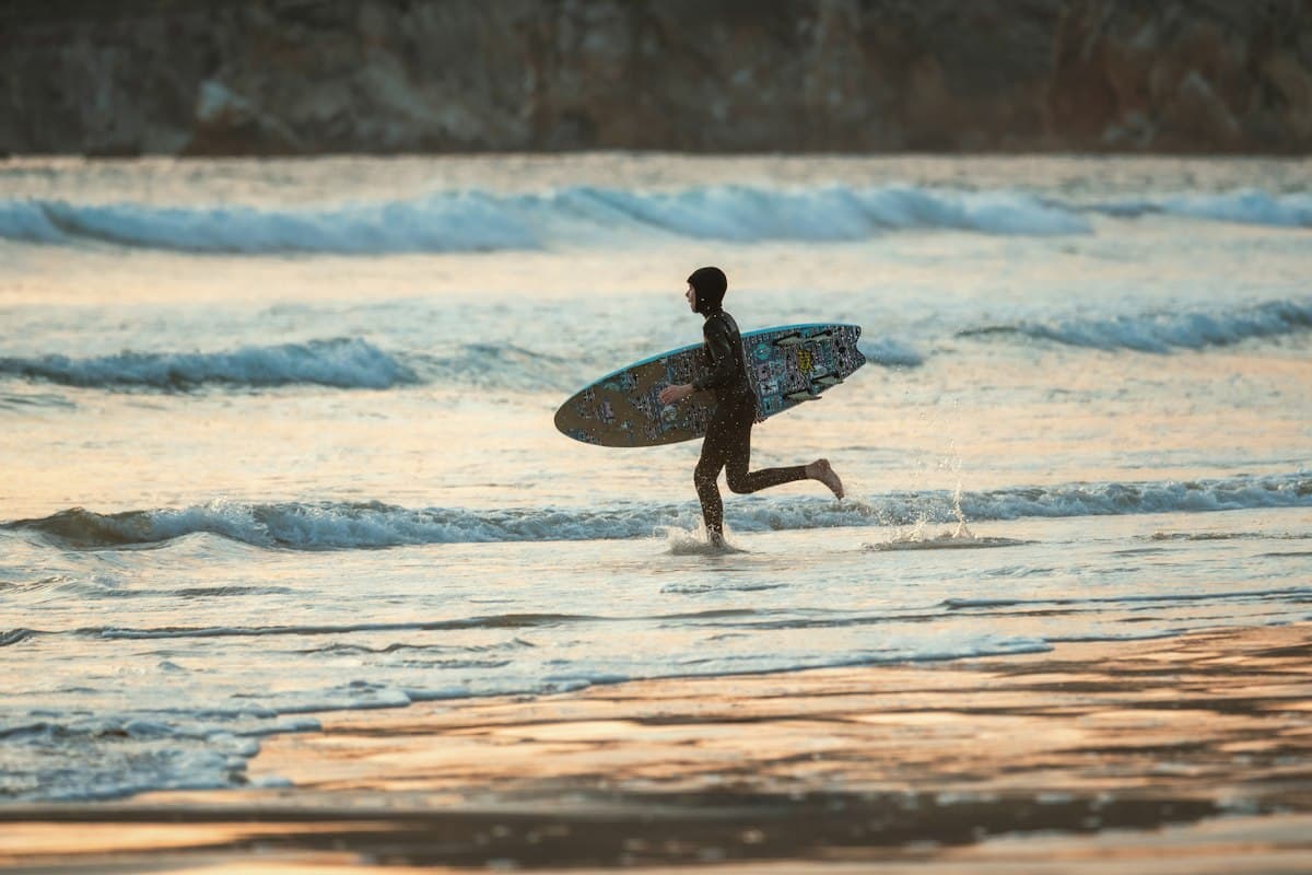 Un surfeur court dans l'écume, sa planche sous le bras, dans la lumière du soir