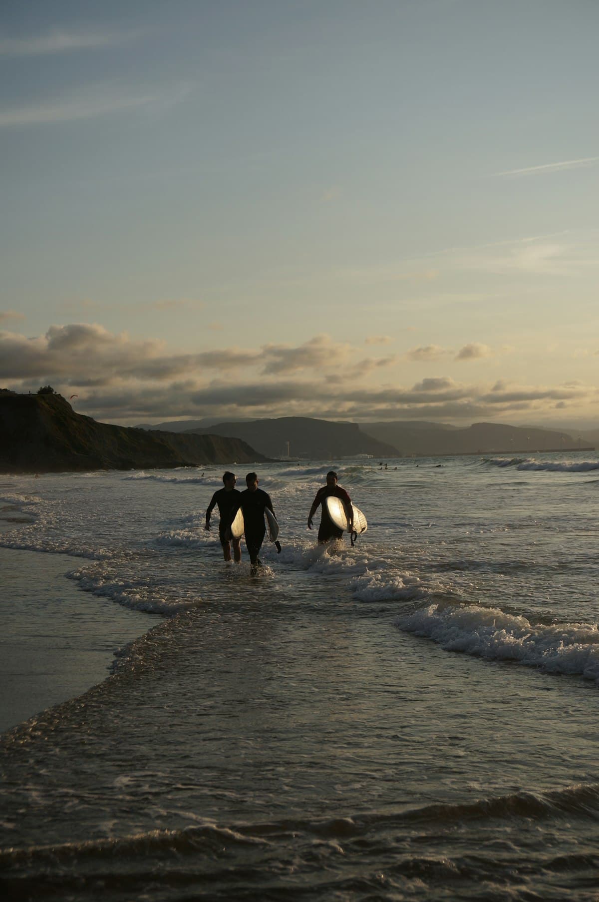Trois surfeurs vus de loin avancent dans l'écume, planches sous le bras