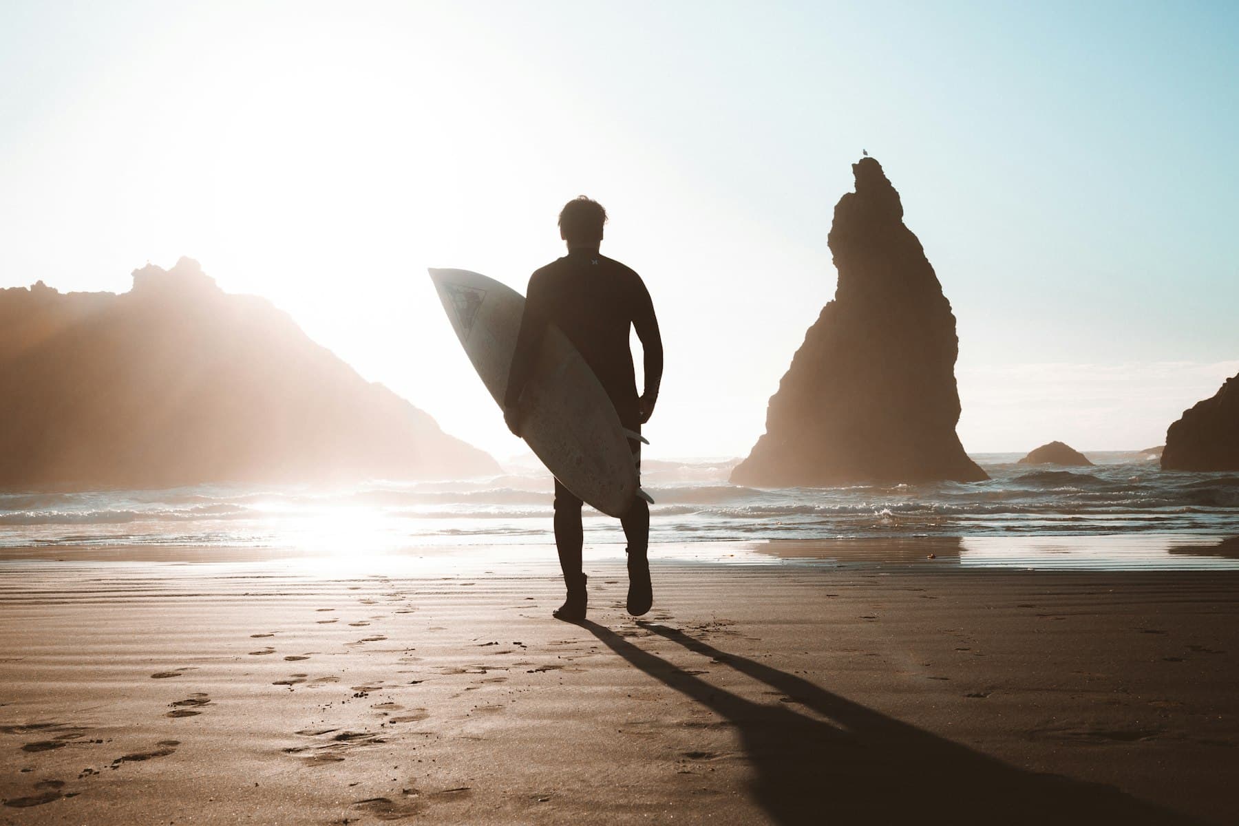Un surfeur en silhouette marche vers l'eau, planche sous le bras, entre deux rochers