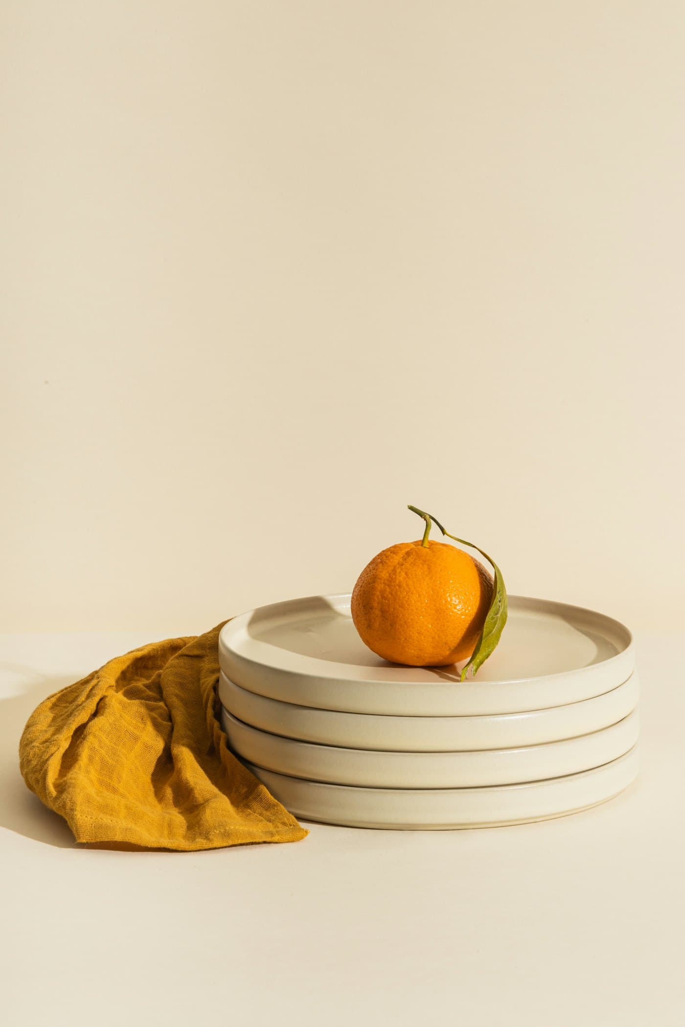 Pile d'assiettes crème avec une clémentine posée dessus, un linge moutarde à côté
