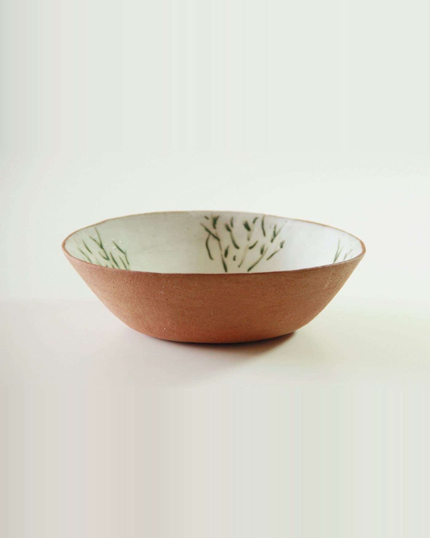 Saladier en terre rouge, intérieur blanc peint d'herbes vertes, sur fond crème