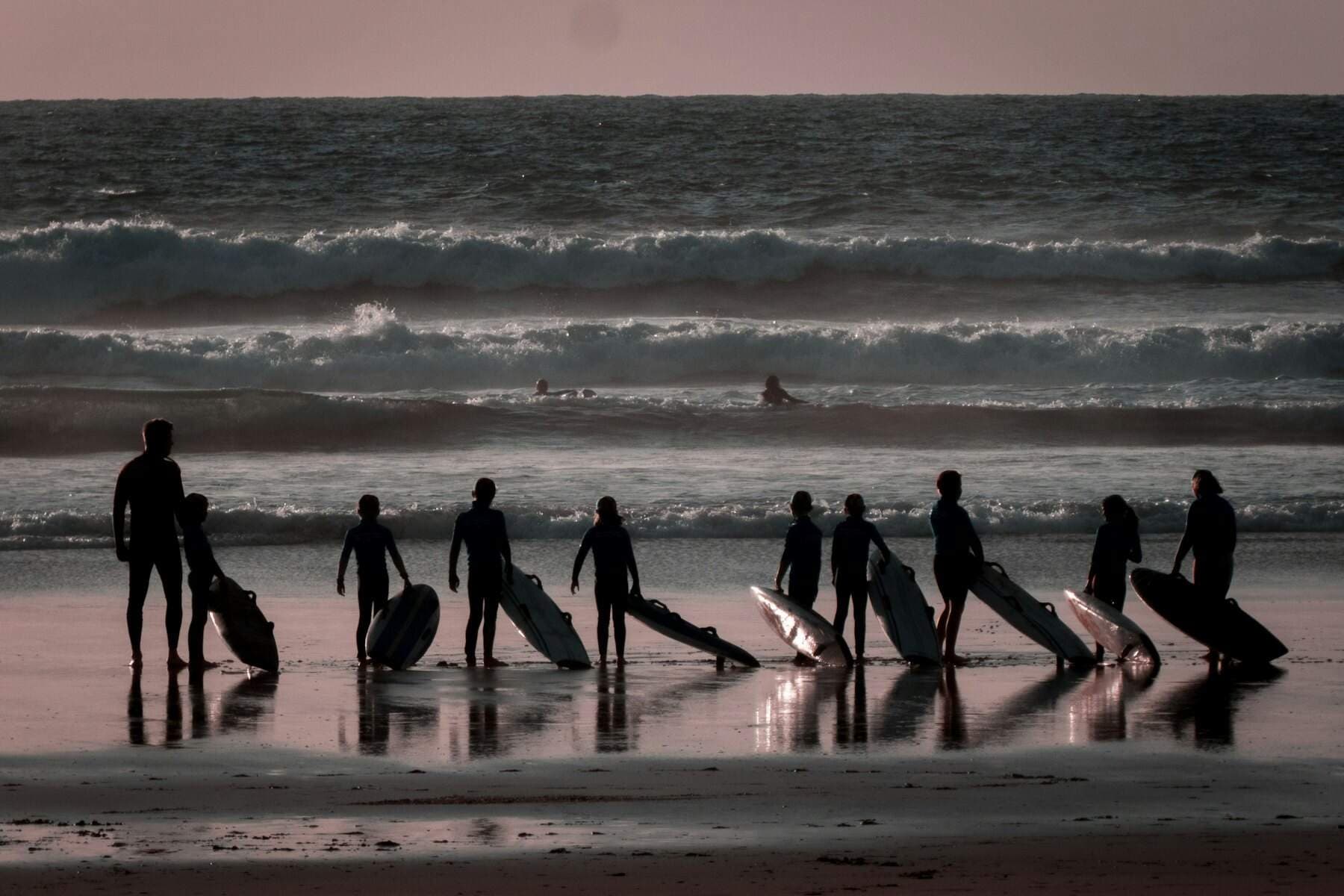 Une dizaine d'élèves en silhouette, planches posées sur le sable mouillé, face aux vagues