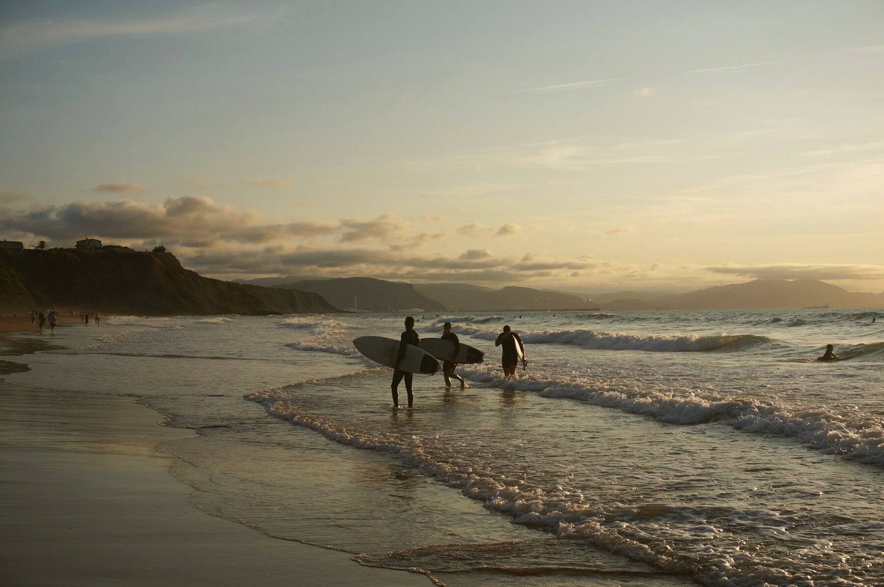 Trois surfeurs entrent dans l'eau à marée basse, au soleil couchant, falaises au loin