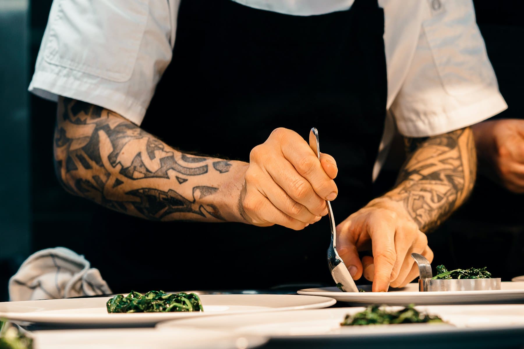 En cuisine, des avant-bras tatoués dressent une assiette à la pince, tablier noir