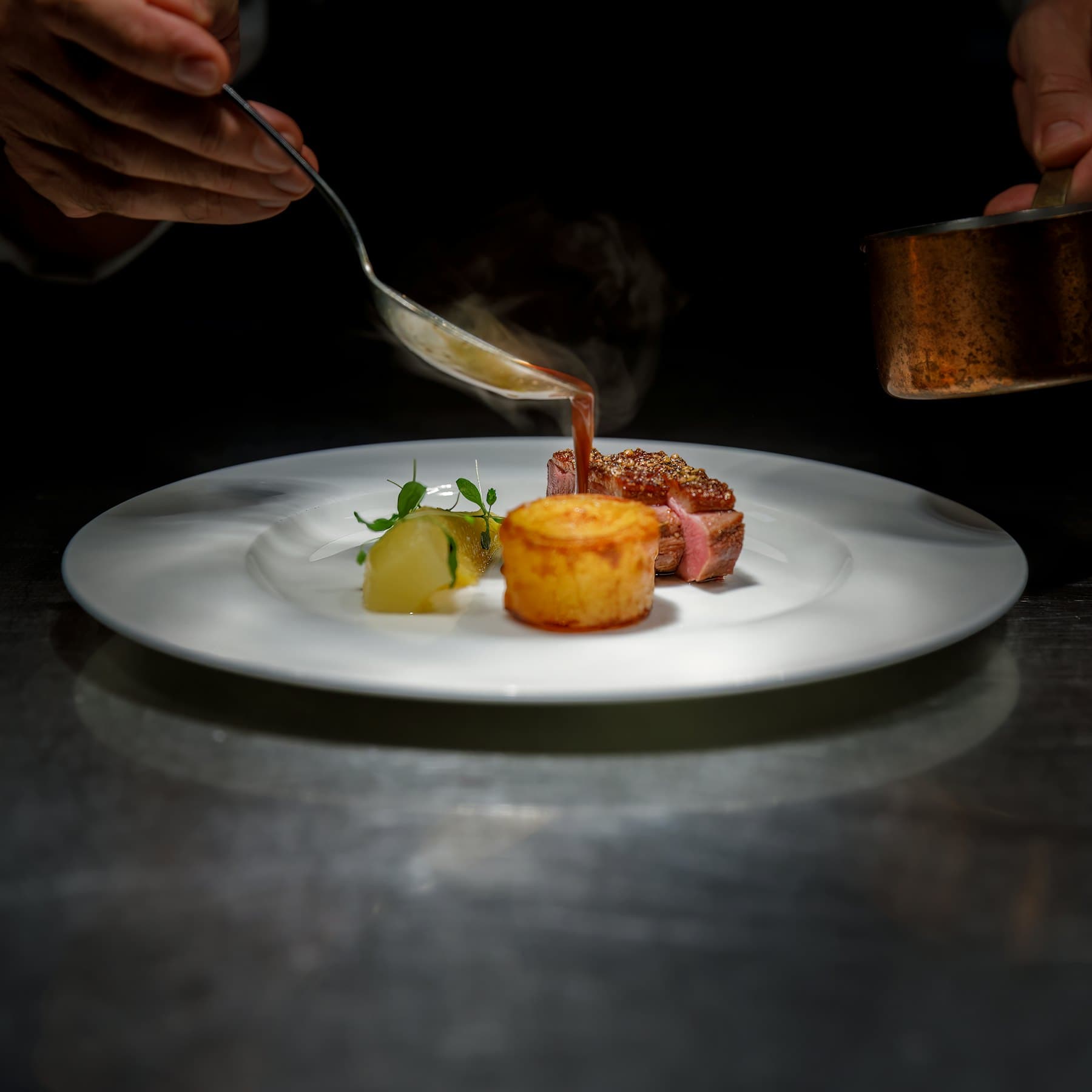 Une cuillère nappe de jus un magret tranché, posé près d'une pomme fondante sur une grande assiette blanche, dans la pénombre