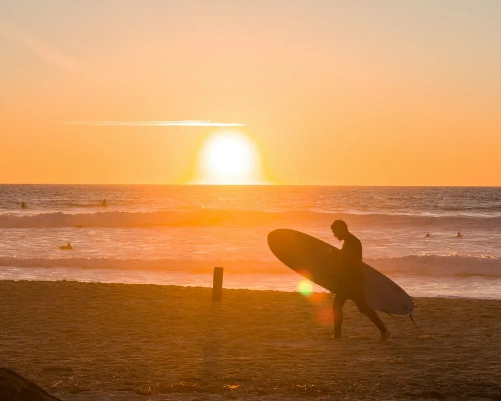 Démonstration fictive : un surfeur marche sur le sable, sa planche sous le bras, face au soleil couchant.